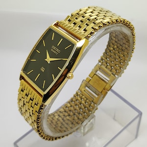 Peut inclure: Montre Seiko Quartz dorée avec un cadran rectangulaire vert foncé. La montre présente des aiguilles et des index dorés, ainsi qu'un bracelet en maille dorée. La montre est présentée sur un présentoir en plastique transparent.