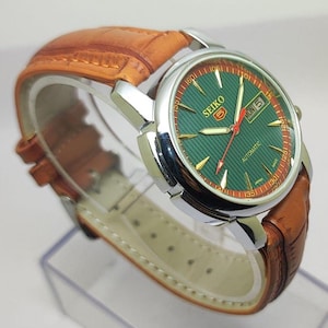 May include: A Seiko 5 automatic wristwatch featuring a round silver-tone stainless steel case, a textured green dial with gold-tone indices, a day-date window at the three o'clock position, and a brown leather strap with a crocodile-embossed pattern.