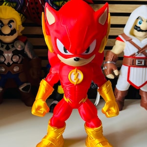 May include: A red and gold figurine depicting a character with spiky hair, large white eyes, and a lightning bolt emblem on the chest. The figure wears gold gauntlets and boots, standing in a forward-facing pose with clenched fists and a serious expression.