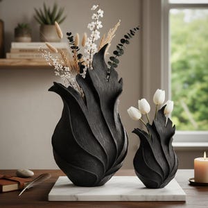May include: Two black, textured vases with a distinctive, flame-like design. One vase holds dried flowers and greenery, the other holds white tulips. The vases sit on a white marble base, with a candle and a notebook in the background.
