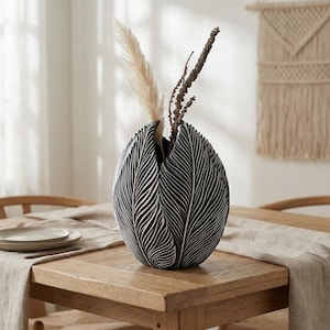 Puede incluir: Un jarrón de cerámica en blanco y negro con un diseño de hojas, que contiene plantas decorativas secas. El jarrón está sobre una mesa de madera con un mantel de color neutro. El jarrón mide unos 25 cm de alto.
