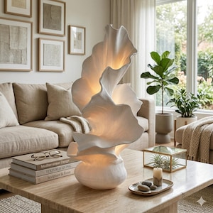 Puede incluir: Una lámpara de mesa escultural blanca con un diseño orgánico en capas. La lámpara tiene una superficie texturizada y emite un brillo cálido. Está sobre una mesa de centro de color claro, junto a libros y objetos decorativos. La lámpara mide unos 76 cm de altura.