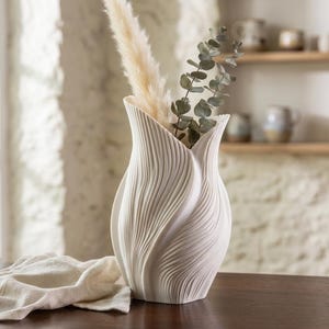 Peut inclure: Un vase en céramique blanc avec un motif texturé en forme de vagues. Le vase contient de l'herbe de la pampa séchée et des branches d'eucalyptus. Décoration d'intérieur. Le vase est posé sur une surface en bois foncé, avec un tissu blanc au premier plan.