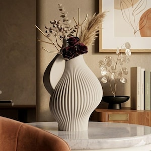May include: A white, ribbed ceramic vase with a unique shape, holding dark red roses, dried grasses, and gold-colored decorative stems. The vase sits on a marble-topped table, showcasing a modern home decor aesthetic.