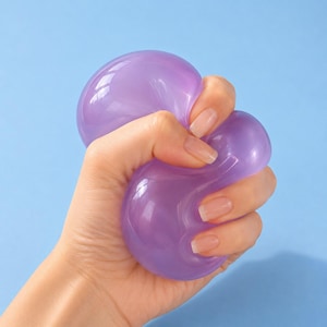 Può includere: Una pallina antistress traslucida, di colore viola chiaro, arrotondata e morbida, realizzata in un materiale liscio e flessibile. L'oggetto viene compresso, mostrando la sua consistenza malleabile e la capacità di cambiare forma.