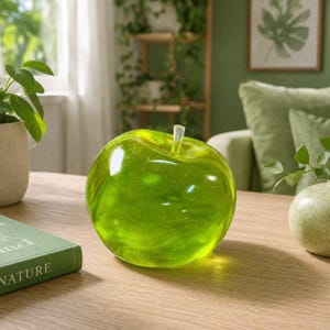 May include: A translucent lime green apple-shaped decorative object made of smooth glass or resin. The object features a rounded, bulbous form with a small, clear, cylindrical stem protruding from the top center, reflecting light on its surface.