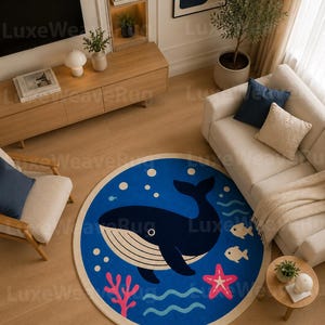 May include: A round area rug featuring a deep blue background with a large dark blue whale in the center. The design includes white bubbles, two small light-colored fish, a pink starfish, and pink coral accents, all bordered by a cream-colored edge.