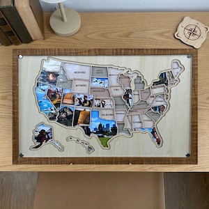 Peut inclure: Une carte encadrée en bois des États-Unis avec des états individuels découpés. Chaque état contient une photographie. Le cadre est marron et le fond est de couleur beige clair. Une petite boussole en bois est dans le coin supérieur droit.