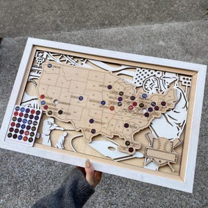 Può includere: Una mappa in legno degli Stati Uniti con ritagli a tema baseball e spilli colorati che segnano le località. La mappa è incorniciata in bianco e include una scheda separata con spilli aggiuntivi. Il design presenta una palla da baseball, una mazza e un giocatore.