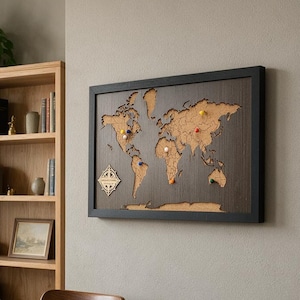 May include: A rectangular wall-mounted world map featuring a dark wood frame and a cork-textured landmass surface. The map includes a decorative compass rose icon in the lower-left corner and several colorful push pins marking various locations.