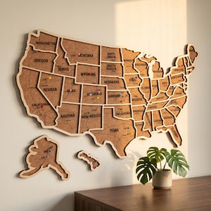 May include: A wall-mounted cork map of the United States featuring individual state cutouts with black printed state names and borders. The map includes Alaska and Hawaii as separate pieces, with colorful push pins inserted into various locations.