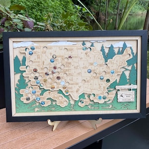 May include: A framed wooden travel map of the United States, featuring a layered design with laser-cut details. The map includes colorful pins marking locations and is surrounded by green trees and a black frame. The sign reads "TRAVEL MAP" with the names "Leo & Luna".