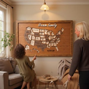 Puede incluir: Un gran mapa de pared rectangular de madera de los Estados Unidos con bordes de estados cortados con láser, nombres de estados grabados e iconos pequeños. El texto "Brown Family" está montado arriba y "USA TRAVEL MAP" abajo.