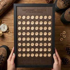 Puede incluir: Una placa de pared rectangular de madera con marco negro, que presenta el texto "NATIONAL PARKS OF THE UNITED STATES" y "EXPLORE AMERICA'S WILDS". Contiene una cuadrícula de recortes circulares, cada uno etiquetado con el nombre de un parque nacional.