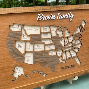 Könnte beinhalten: Eine USA-Reisekarte aus Holz mit dem Text Brown Family oben und USA TRAVEL MAP ADVENTURE AWAITS unten. Die Karte besteht aus einzelnen Bundesstaat-Teilen in hellen und dunklen Holztönen, die jeweils mit Staatsnamen und Symbolen graviert sind.