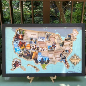 Può includere: Una mappa da parete incorniciata degli Stati Uniti con sagome in legno dei singoli stati riempite di fotografie personali. La mappa include una rosa dei venti nell'angolo in basso a destra con il testo Robinson's Family USA Travel Map.