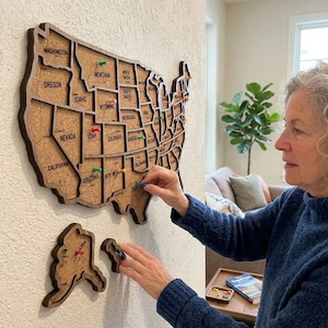 May include: A wall-mounted cork map of the United States featuring dark wood borders and individual state cutouts. The map includes Alaska and Hawaii as separate pieces, with colorful push pins inserted into various states to mark locations.