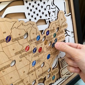 May include: A layered wooden map of the United States featuring laser-cut state borders and stadium names. Small, colorful football-shaped markers are placed on the map, representing various professional football stadiums across the country.