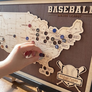 May include: A wooden wall map of the United States featuring the text BASEBALL MAJOR LEAGUE and Michael Family Stadium Map. The map includes circular cutouts for placing team logo markers, with several markers already positioned on the board.