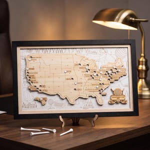 May include: A framed wooden map of the United States featuring laser-cut state borders and golf course locations marked with white pins. The map includes a decorative emblem with a golf ball and the text "TOMMY'S GOLF TOUR MAP" in a black frame.