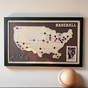 May include: A framed wooden map of the United States featuring the text BASEBALL MAJOR LEAGUE and Athletics. The map includes circular markers on various states and a small rectangular photo frame labeled SMITH FAMILY. A baseball sits below the frame.