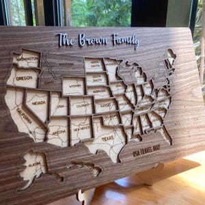 May include: A wooden wall map of the United States featuring laser-cut state borders and names. The text "The Brown Family" is displayed at the top, and "USA TRAVEL MAP ADVENTURE AWAITS" is printed in the bottom right corner with a compass rose.