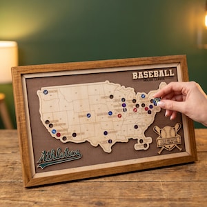 May include: A framed wooden map of the United States featuring state outlines and circular cutouts for baseball stadium tokens. The text BASEBALL MAJOR LEAGUE is at the top, with Athletics and a baseball graphic at the bottom of the brown board.