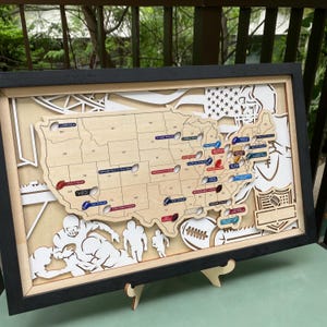 Puede incluir: Un mapa de madera enmarcado de los Estados Unidos con fronteras estatales cortadas con láser y pequeños marcadores de colores que representan estadios de la NFL. El fondo incluye recortes blancos superpuestos de jugadores, un balón y una bandera.