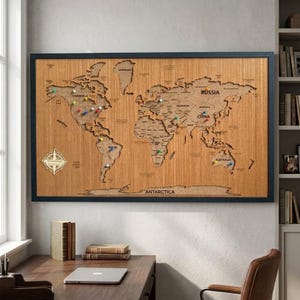 May include: A rectangular wooden world map wall art piece framed in black. The map features laser-cut continents with engraved country names and borders. Several colorful push pins are inserted into various locations. A compass rose is visible.