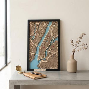 Könnte beinhalten: Eine gerahmte Holzkarte von Manhattan, New York City, in einem schwarzen Rahmen. Die Karte hat ein detailliertes, geschichtetes Design mit Braun- und Beigetönen, blauem Wasser und roten Markierungen. Der Text "NEW YORK DREAMIN" ist sichtbar.