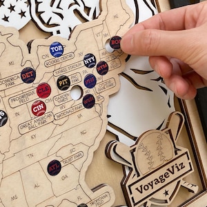 Może przedstawiać: Drewniana mapa stadionów baseballowych z przypinkami o tematyce baseballowej. Mapa przedstawia nazwy i lokalizacje stadionów w Stanach Zjednoczonych. Przypinki są okrągłe z logo drużyn. Widoczne jest logo VoyageViz.