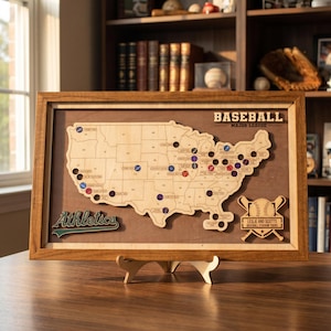 May include: A framed wooden map of the United States featuring a laser-cut design with the text BASEBALL MAJOR LEAGUE, Athletics, and LESLIE AND SCOTTS BASEBALL STADIUM TOURS. The map includes circular cutouts and coloured markers for stadium locations.