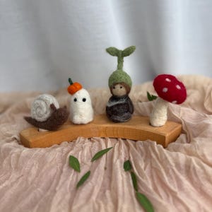 Könnte beinhalten: Ein Set aus vier nadelgefilzten Figuren auf einem geschwungenen Holzständer. Die Figuren bestehen aus einer braunen Schnecke mit weißem Gehäuse, einem weißen Geist mit orangefarbenem Kürbishut, einer dunklen Figur mit grünem Sprossenhut und einem roten Pilz.