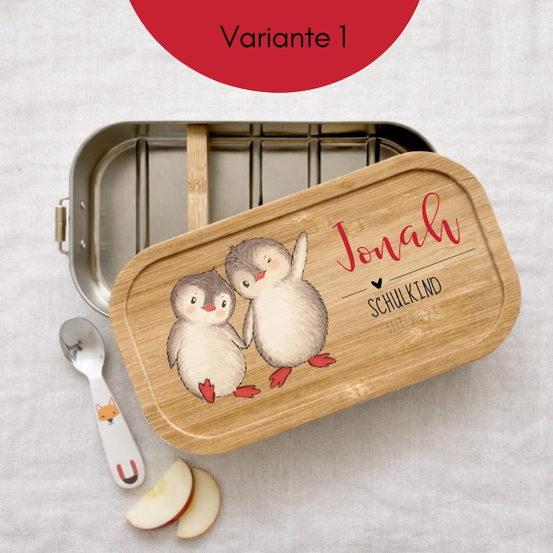 May include: A rectangular stainless steel lunch box with a bamboo lid featuring a printed illustration of two penguins, the name Jonah, the text SCHULKIND, and the year 2024. A small metal spoon with a fox graphic and two apple slices are included.