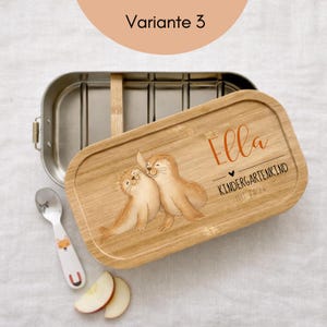 May include: A rectangular stainless steel lunch box with a removable bamboo lid featuring a printed illustration of two seals, the name Ella, and the text KINDERGARTENKIND SHI 2024. A small metal spoon with a fox graphic and two apple slices are included.