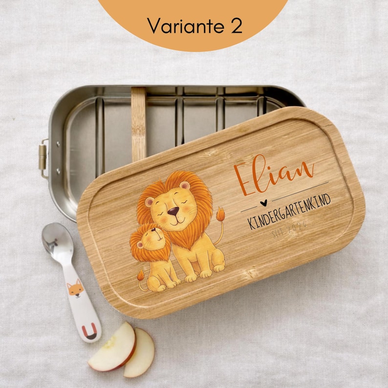 May include: A rectangular stainless steel lunch box with a bamboo lid featuring a printed illustration of two lions, the name Elian, and the text KINDERGARTENKIND SEIT 2024. A small silver spoon with a fox graphic and two apple slices are nearby.