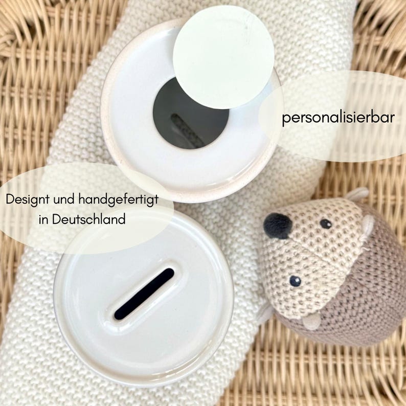May include: A white ceramic piggy bank with a coin slot, next to a knitted hedgehog toy. The piggy bank is designed and handcrafted in Germany. The text "personalizable" is also visible.