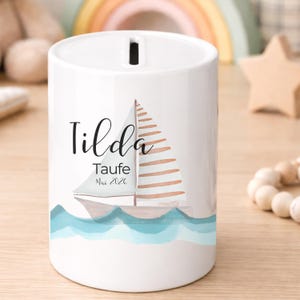 May include: White ceramic bank with a sailboat design and the name "Tilda". The sailboat has a white sail and brown stripes. The bank has a slot at the top for inserting money and blue waves at the bottom.