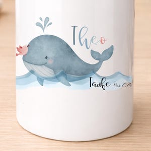 May include: White ceramic mug featuring a watercolor illustration of a blue whale with a pink bird. The mug has the name "Theo" in blue script and the word "Taufe" with the date "Mai 2026".