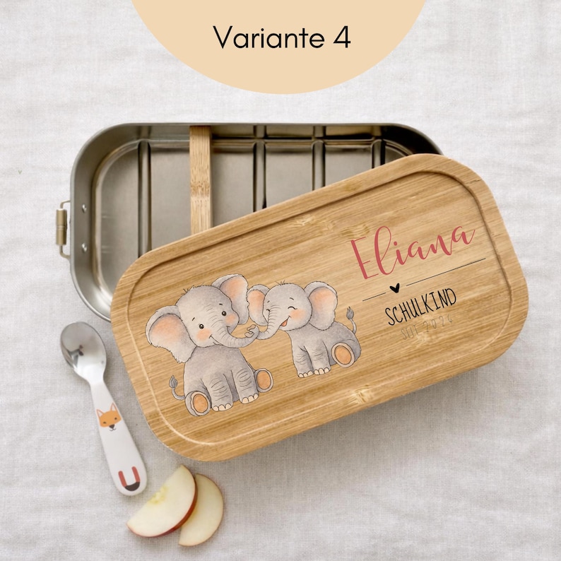 May include: A rectangular stainless steel lunch box with a bamboo lid featuring two illustrated elephants, the name Eliana, and the text SCHULKIND SEIT 2026. A small silver spoon with a fox graphic and two apple slices are placed nearby.