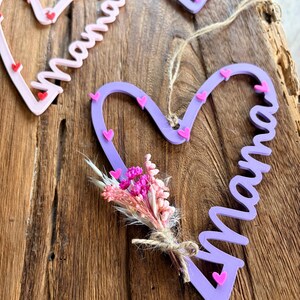 May include: Heart-shaped wooden decorations with the word "Mama" in cursive. The hearts are in shades of pink and purple, decorated with small pink hearts. One heart has a small bouquet of dried flowers.
