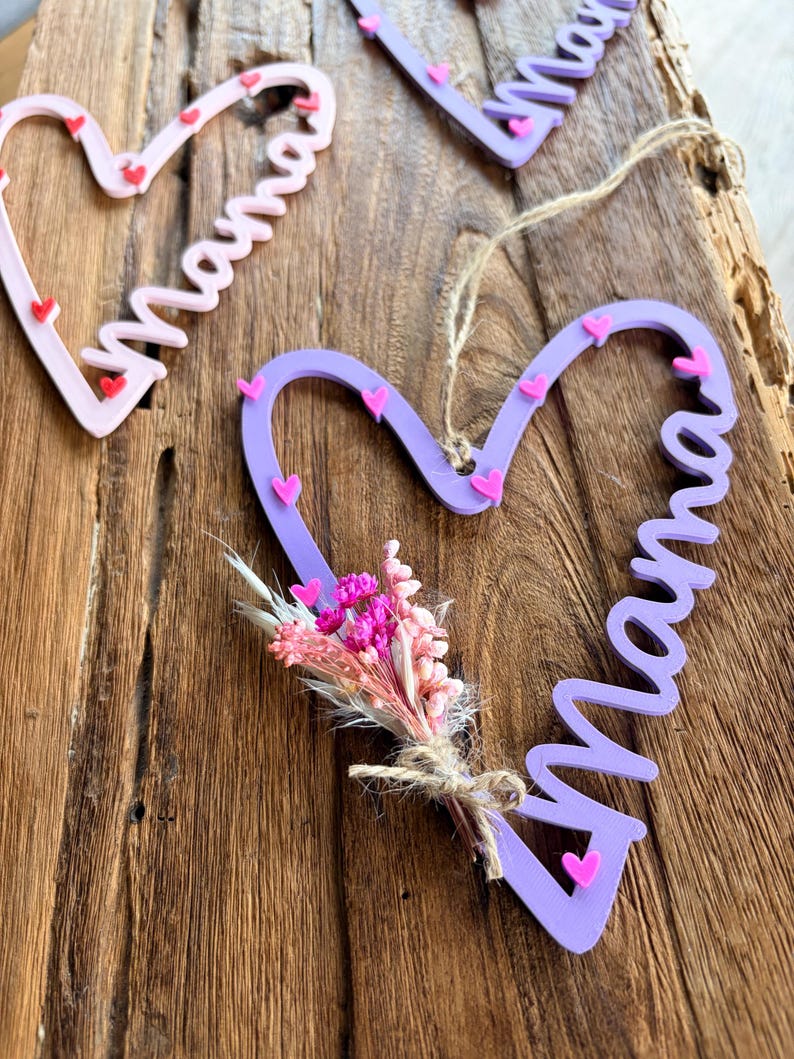May include: Heart-shaped wooden decorations with the word "Mama" in cursive. The hearts are in shades of pink and purple, decorated with small pink hearts. One heart has a small bouquet of dried flowers.