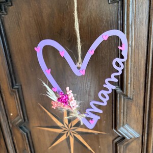 May include: A lavender heart-shaped wall hanging with the word "Mama" in cursive. The heart is decorated with small pink hearts and a small bouquet of dried flowers. A natural twine is used for hanging.