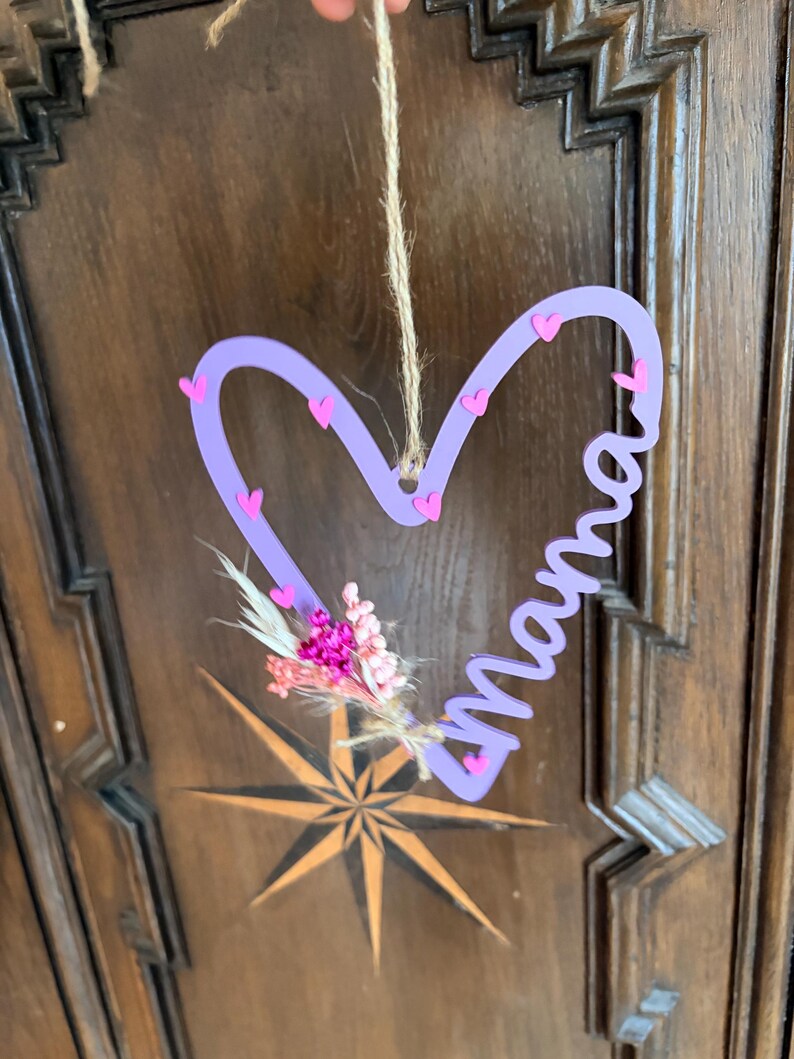 May include: A lavender heart-shaped wall hanging with the word "Mama" in cursive. The heart is decorated with small pink hearts and a small bouquet of dried flowers. A natural twine is used for hanging.