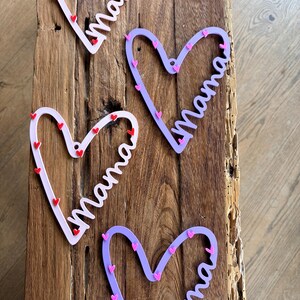 May include: Four heart-shaped decorations with the word "Mama" in a cursive font. Two are light pink with red hearts, and two are purple with pink hearts. The decorations are on a wooden surface.
