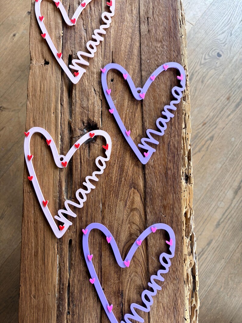 May include: Four heart-shaped decorations with the word "Mama" in a cursive font. Two are light pink with red hearts, and two are purple with pink hearts. The decorations are on a wooden surface.