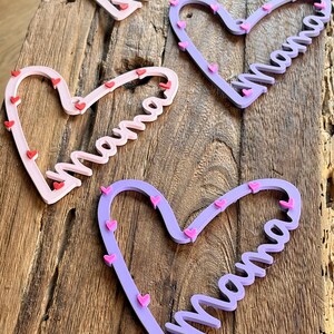 May include: Four heart-shaped wooden decorations with the word "Mama" in cursive. The hearts are in shades of pink and purple, adorned with small red and pink heart accents. They are arranged on a rustic wooden surface.