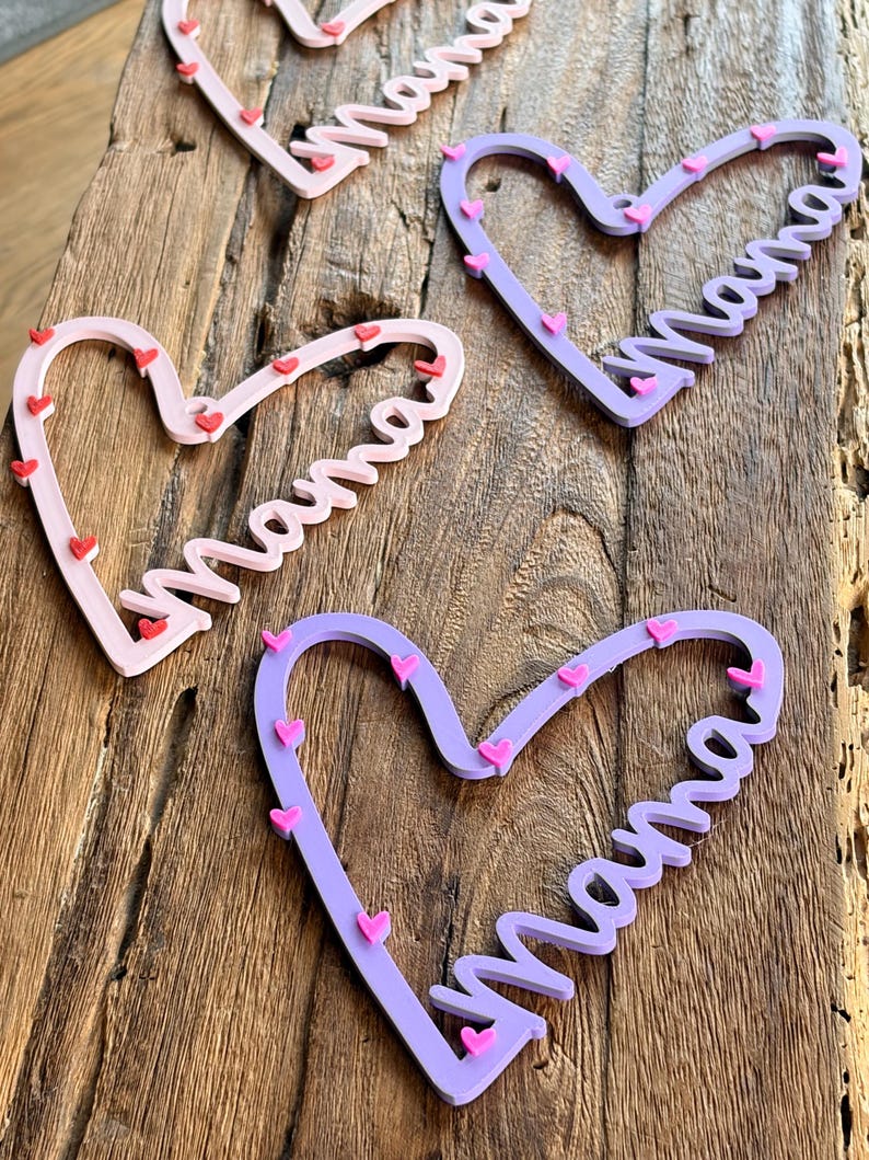 May include: Four heart-shaped wooden decorations with the word "Mama" in cursive. The hearts are in shades of pink and purple, adorned with small red and pink heart accents. They are arranged on a rustic wooden surface.