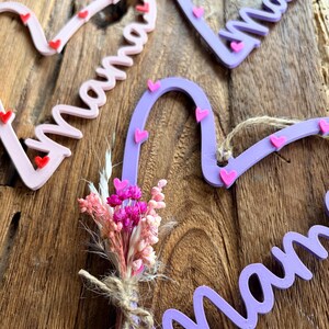 May include: Heart-shaped wooden decorations spelling "Mama" in cursive, decorated with small pink and red hearts. A small bouquet of dried flowers is tied with twine. The items are in shades of pink and purple.