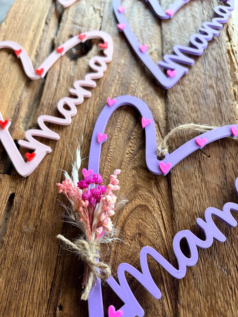 May include: Heart-shaped wooden decorations spelling "Mama" in cursive, decorated with small pink and red hearts. A small bouquet of dried flowers is tied with twine. The items are in shades of pink and purple.